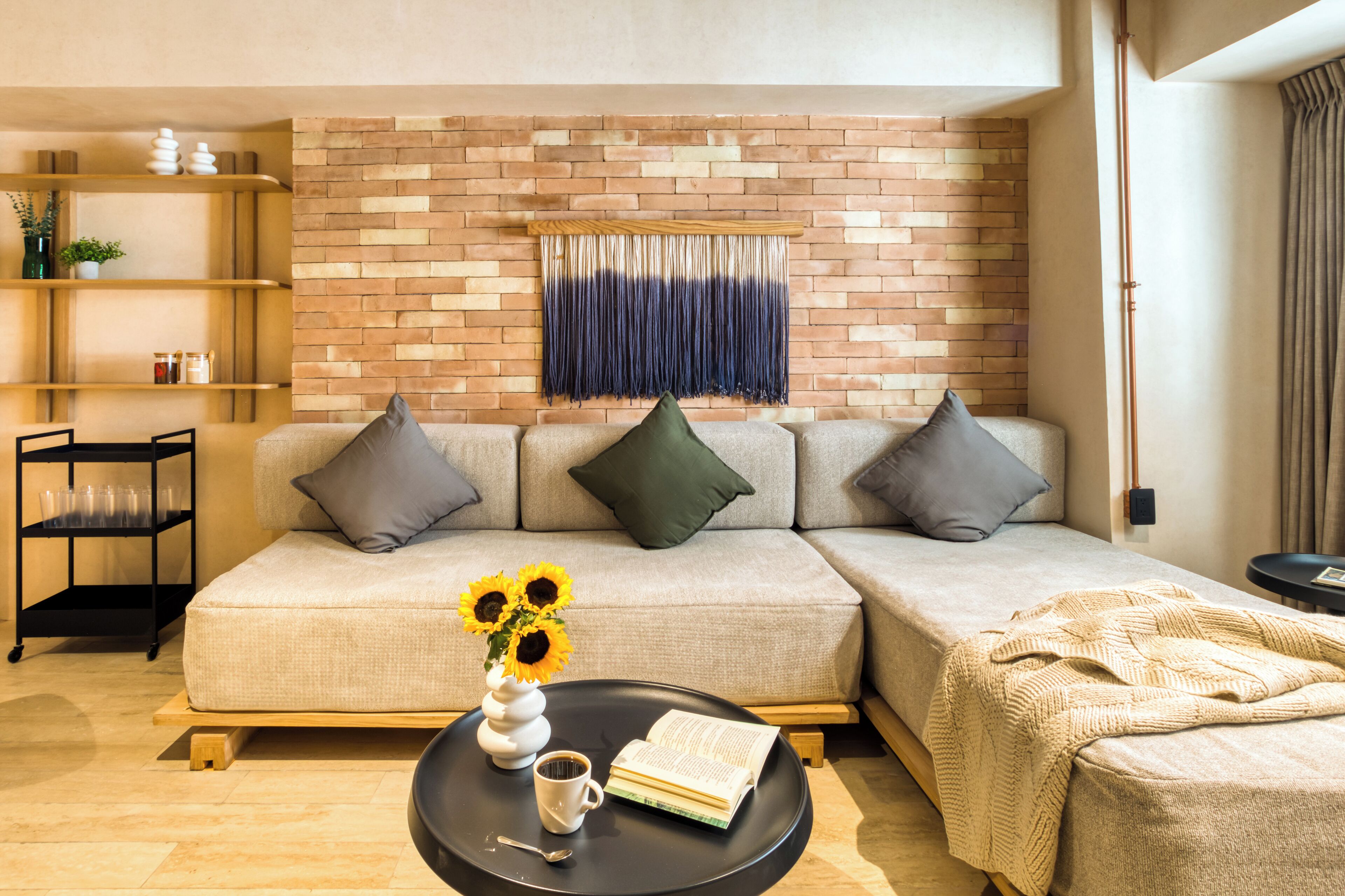 Ein modernes Wohnzimmer mit Backsteinwand, einer Couch, einem Couchtisch mit einem Buch und einer Vase mit Sonnenblumen sowie einem Beistelltisch mit einer Decke.