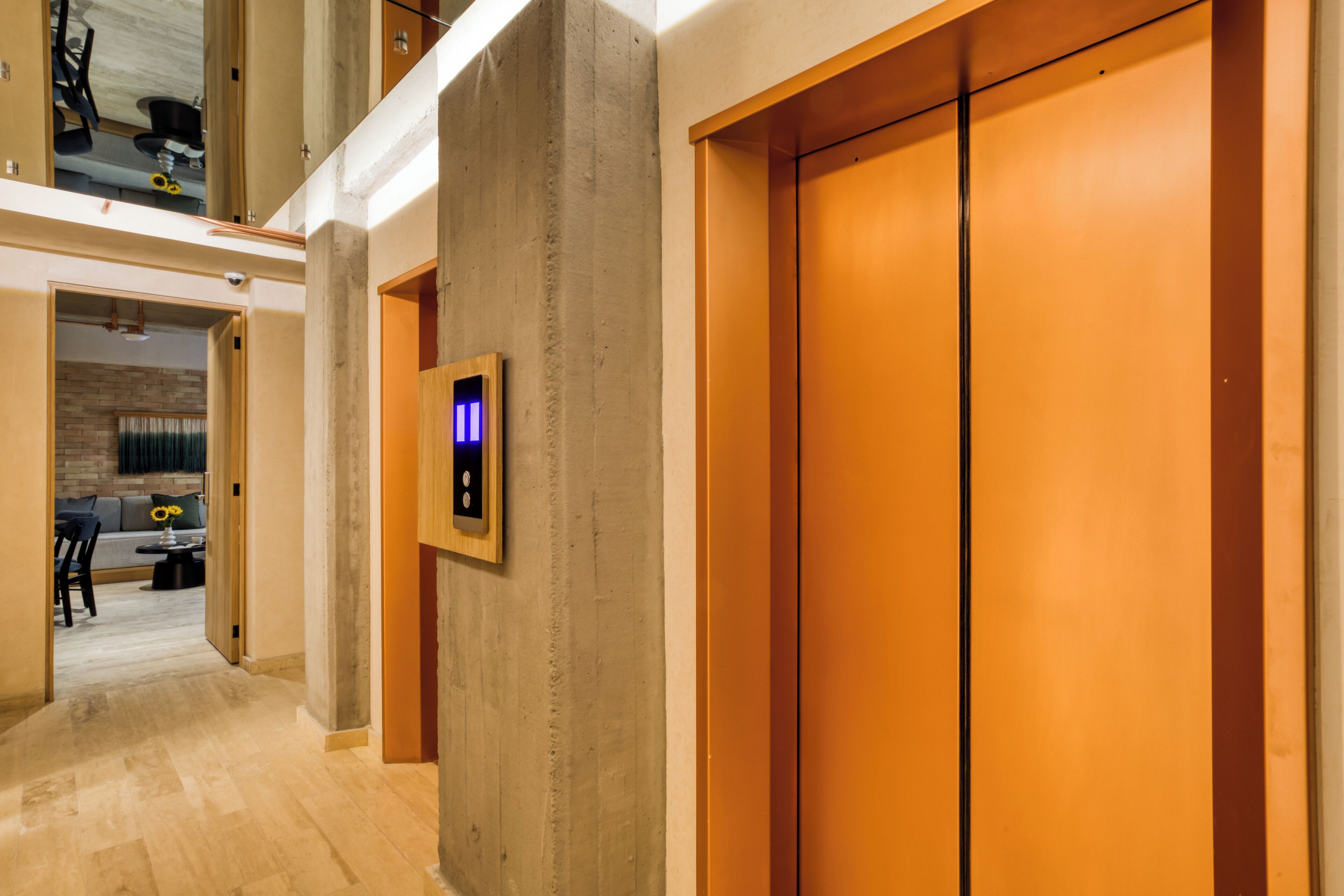 A modern elevator with a wooden finish, a visible emergency call button, and a hallway leading to a seating area with a brick wall and a sofa.
