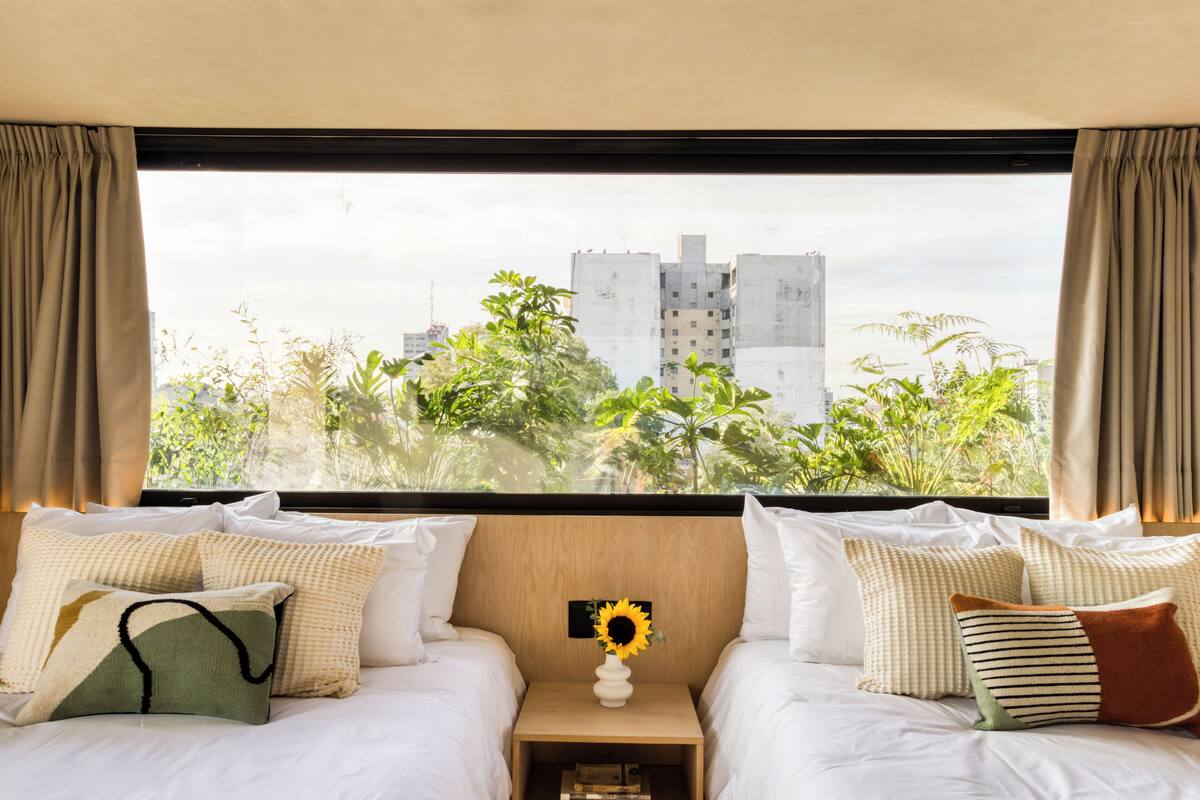 A hotel room with two beds, a nightstand with a vase of sunflowers, and a view of buildings and greenery outside.