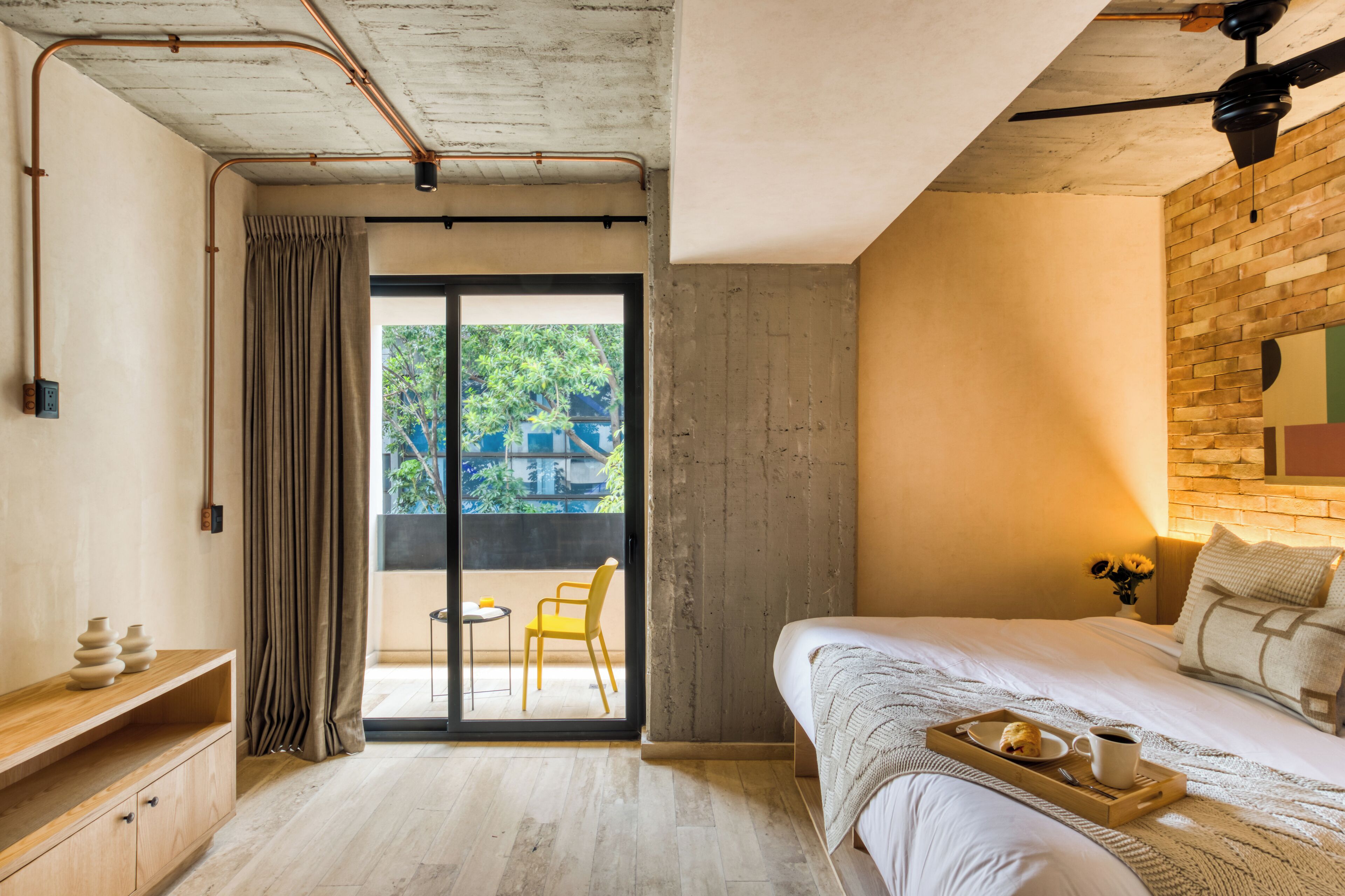A modern bedroom with a large bed, a wooden nightstand, a sliding glass door leading to a balcony, and a brick accent wall.