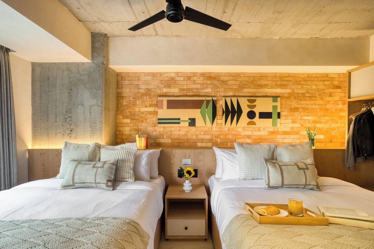 A modern hotel room with two beds, a wooden headboard, a brick accent wall, and a ceiling fan.