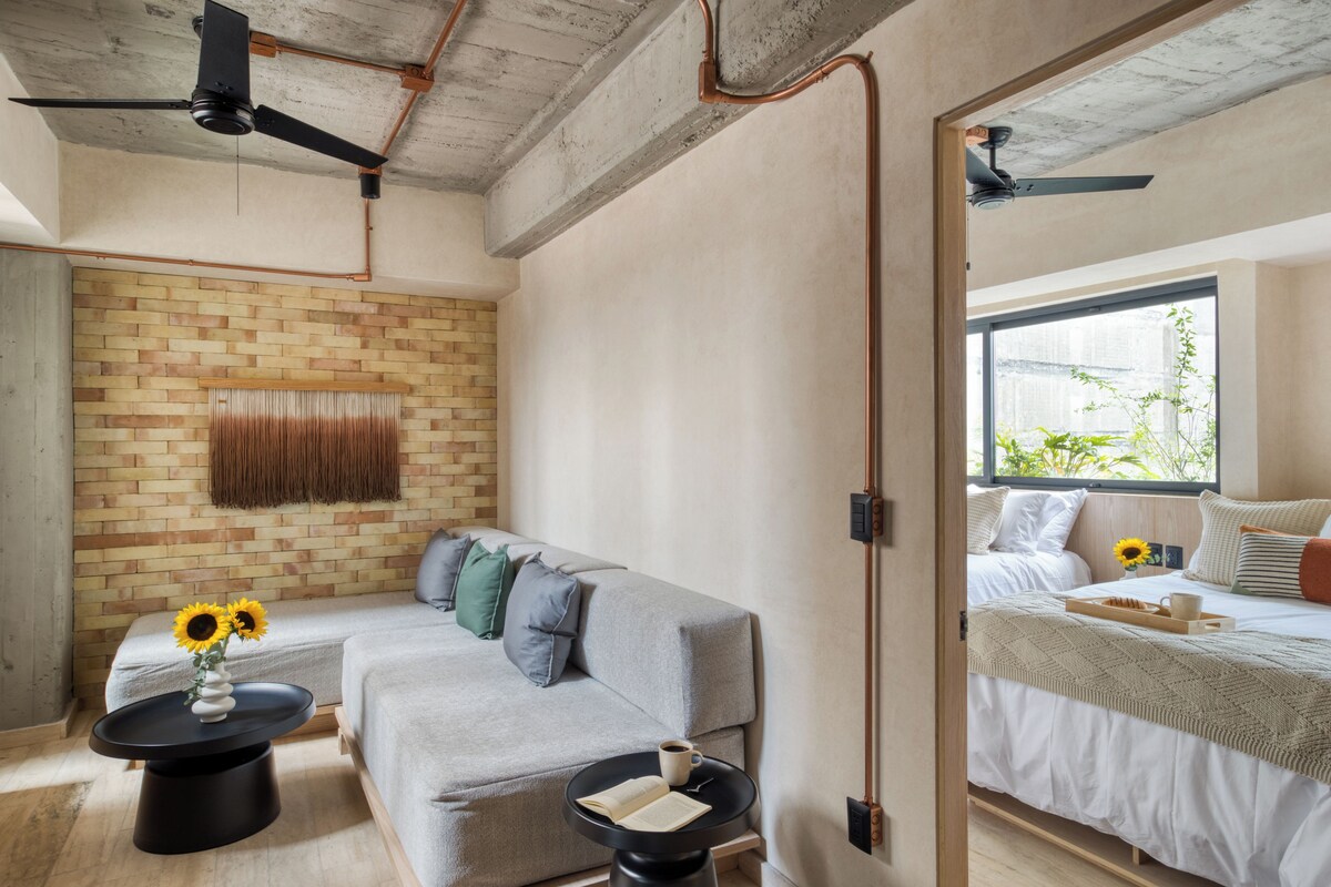 Una habitación moderna con pared de ladrillo, sofá gris, una mesita redonda con un jarrón de girasoles y una cama con vista al exterior.