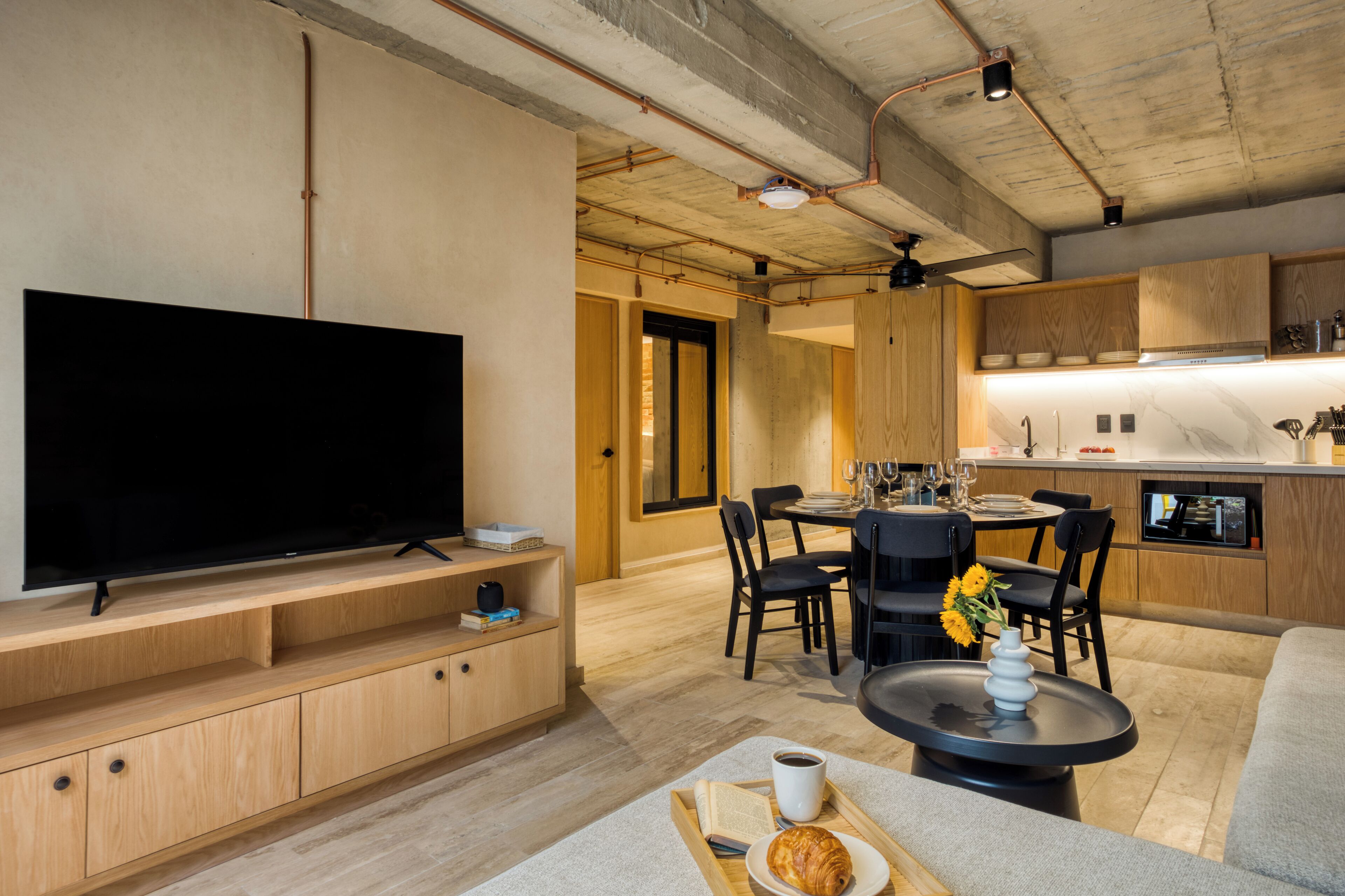 Un salon moderne avec un téléviseur à écran plat posé sur un meuble en bois, un coin repas avec une table ronde et des chaises, et une cuisine dotée de meubles en bois et d’un plan de travail en marbre.
