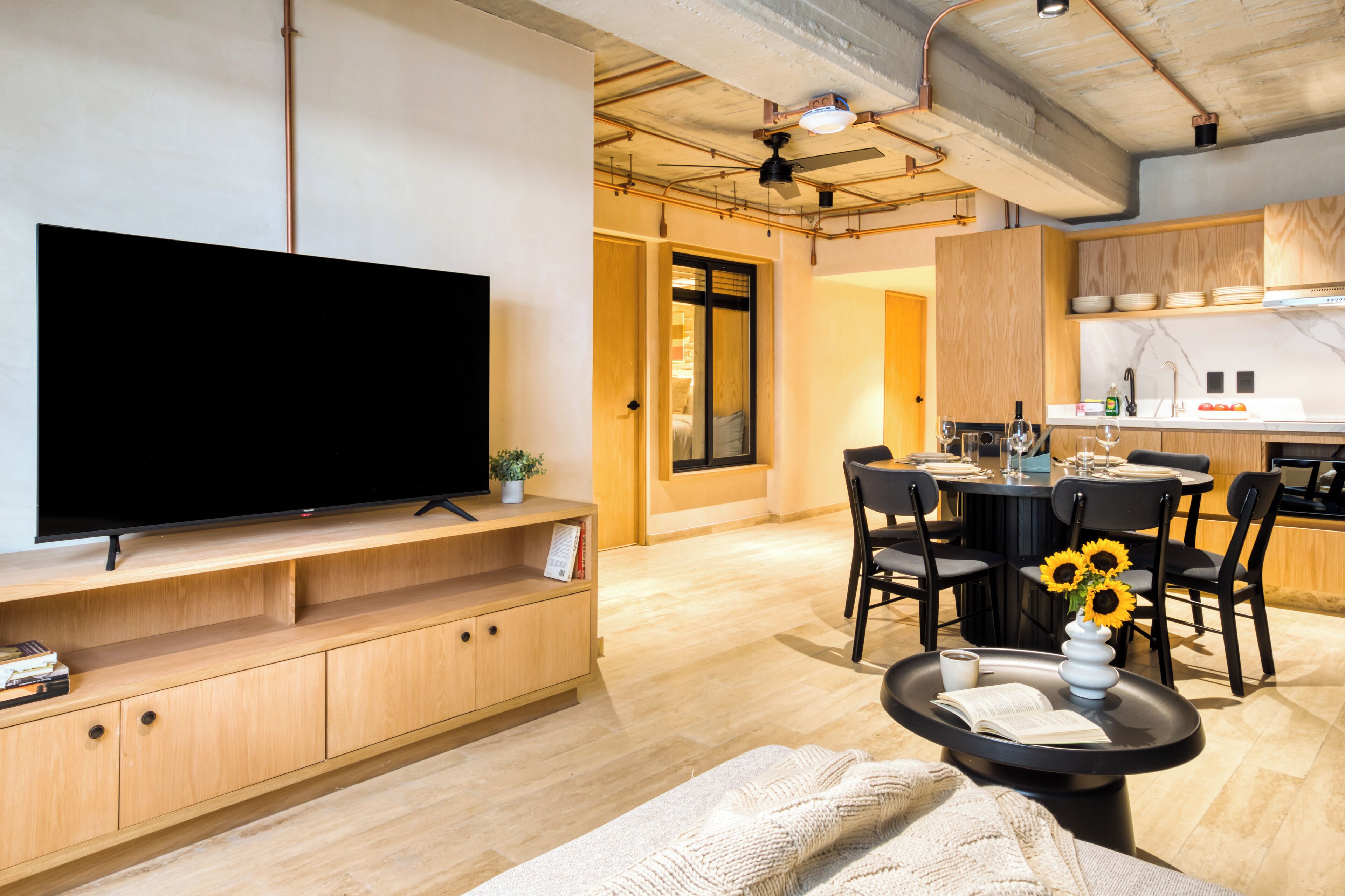 A modern living room with a flat-screen TV, wooden cabinets, a dining area with a round table and chairs, and a kitchen area with wooden cabinets and a white countertop.