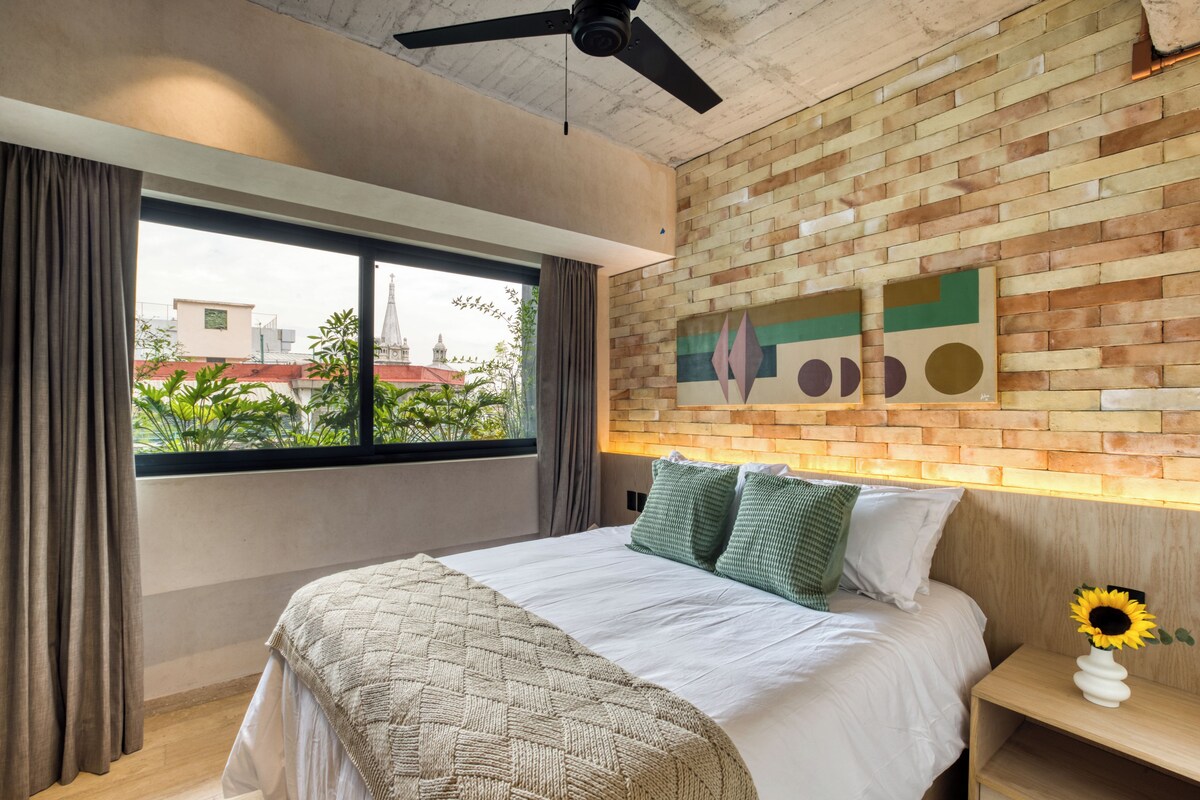 A modern hotel room with a large bed, a view of the cityscape, a brick accent wall, and a ceiling fan.