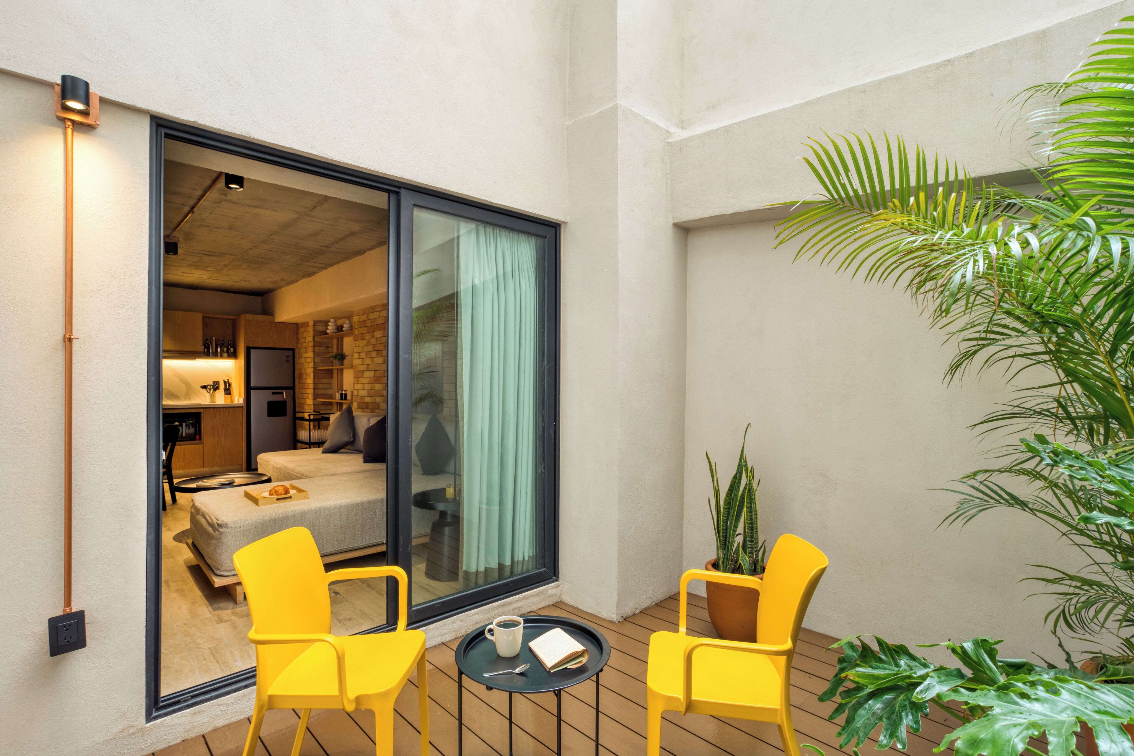 A modern hotel room with a sliding glass door leading to a balcony, yellow chairs, a small table with a cup and book, and a large potted plant.