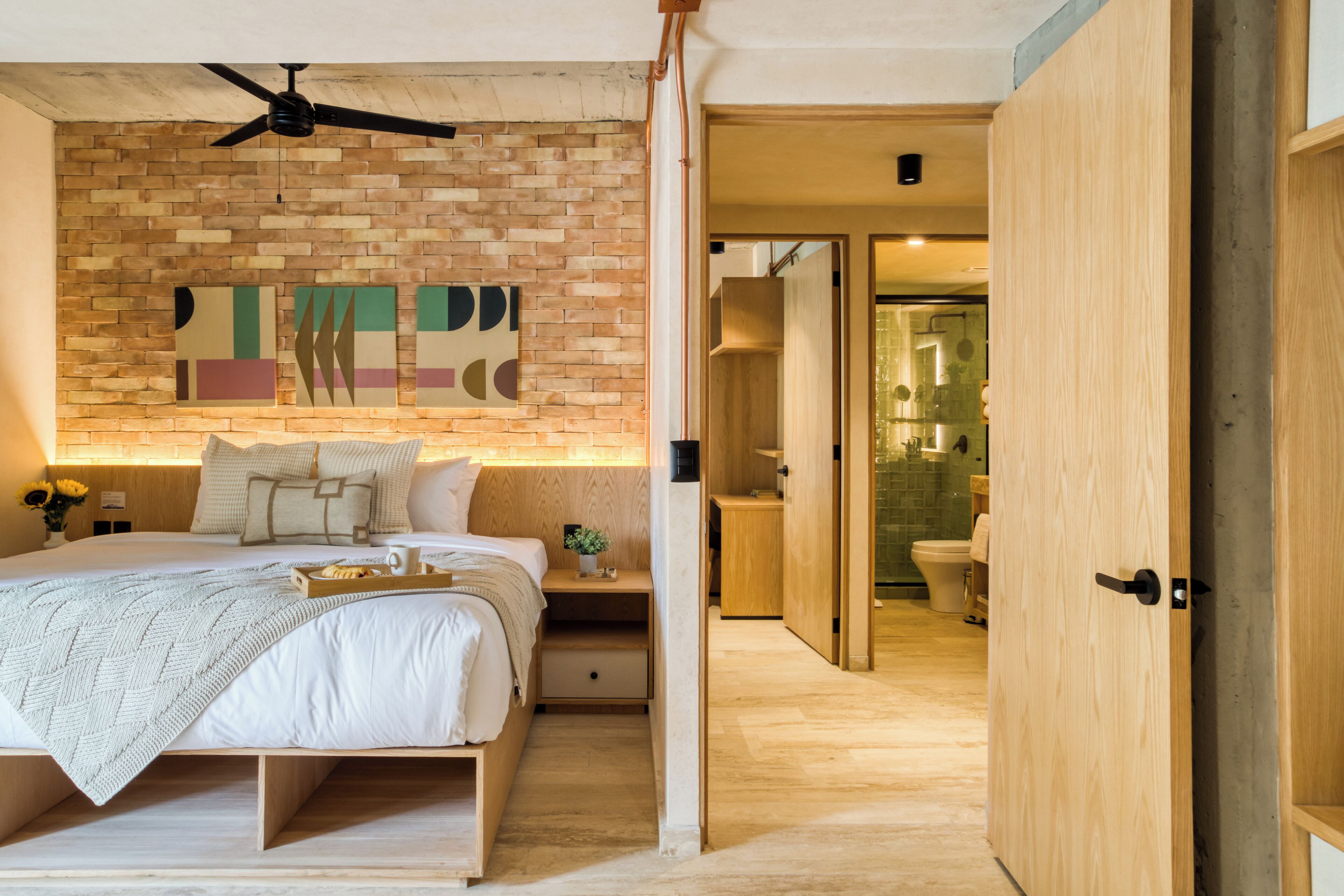 Un dormitorio moderno con pared de ladrillo, ventilador de techo, cama con una bandeja de objetos y armario de madera.