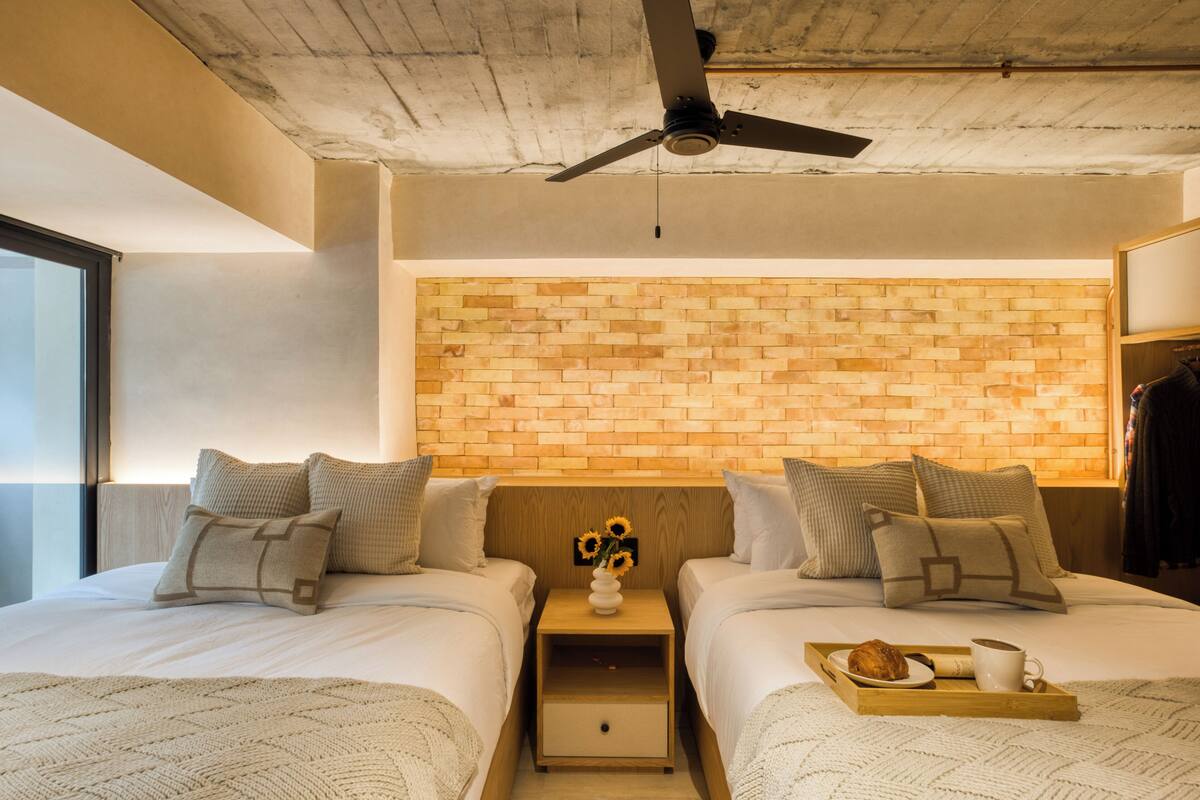 A modern hotel room with two beds, a brick-patterned headboard, a ceiling fan, and a tray with breakfast items.