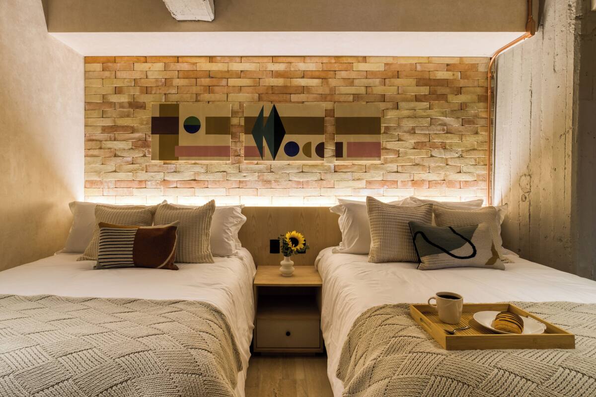 A double room with two beds, a brick-patterned headboard, a nightstand with a vase of sunflowers, and a tray with a cup and a pastry.