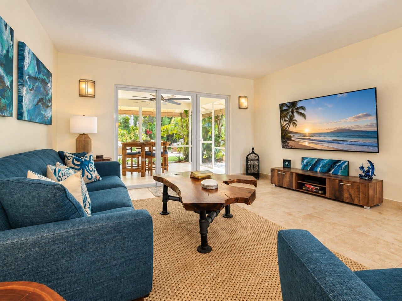 Een moderne woonkamer met een blauwe bank, een houten salontafel en een grote televisiescherm waarop een strand te zien is.