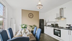 Una cucina moderna con mobili bianchi, una cappa in acciaio inossidabile e un forno a incasso. C'è un'area pranzo con un tavolo apparecchiato per quattro, sedie blu e un lampadario sopra.