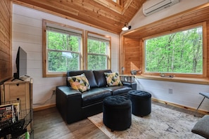 A cozy cabin interior with a black leather sofa, two ottomans, a wooden TV stand, and large windows offering a forest view. - Cozy cabin in charming Cosby with AC (Cosby)