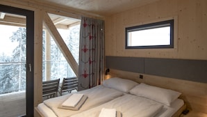 A wooden cabin bedroom with a bed, two pillows, a nightstand, a window, and a view of snow-covered trees.