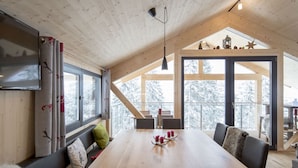 A cozy cabin interior with a wooden dining table, benches, and a large window offering a view of snow-covered trees.