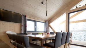 A cozy cabin interior with a wooden dining table, four chairs, a sofa, and a TV mounted on the wall.