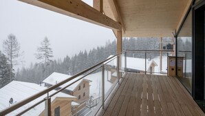 Ein Holzbalkon mit Glasgeländer, der auf eine verschneite Landschaft mit Gebäuden blickt.