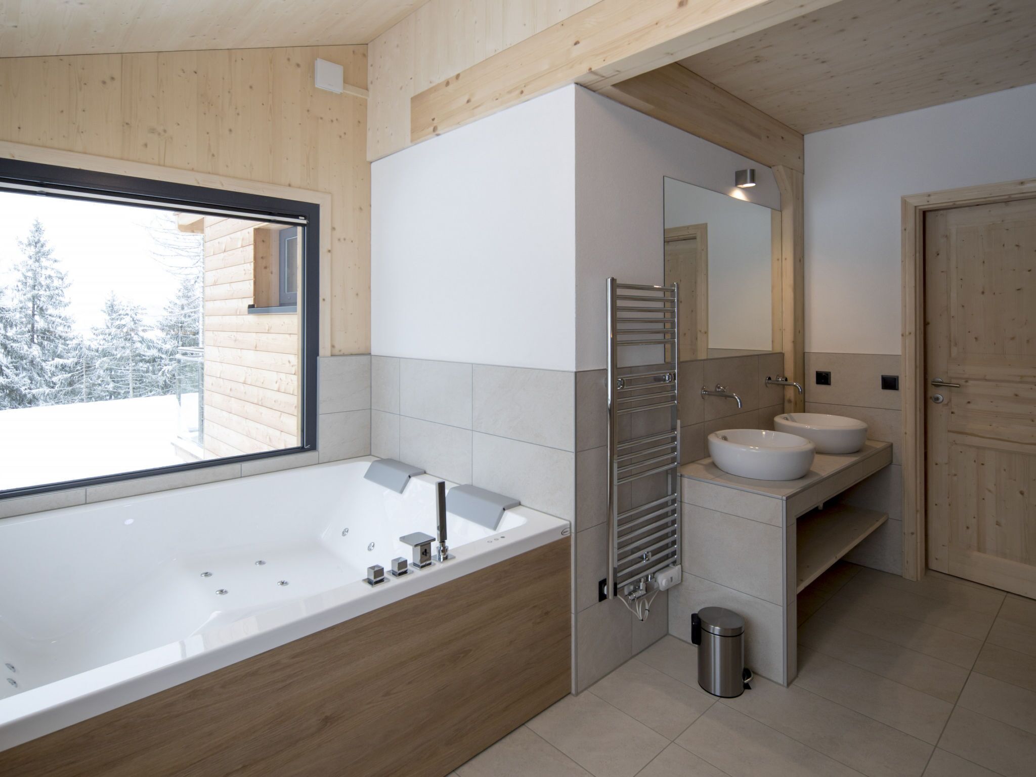A modern bathroom with a large bathtub, two sinks, a mirror, and a window overlooking a snowy landscape.