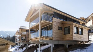 Un bâtiment moderne en bois, à plusieurs étages, doté de balcons et de grandes fenêtres, situé sur une pente enneigée avec des montagnes en arrière-plan.