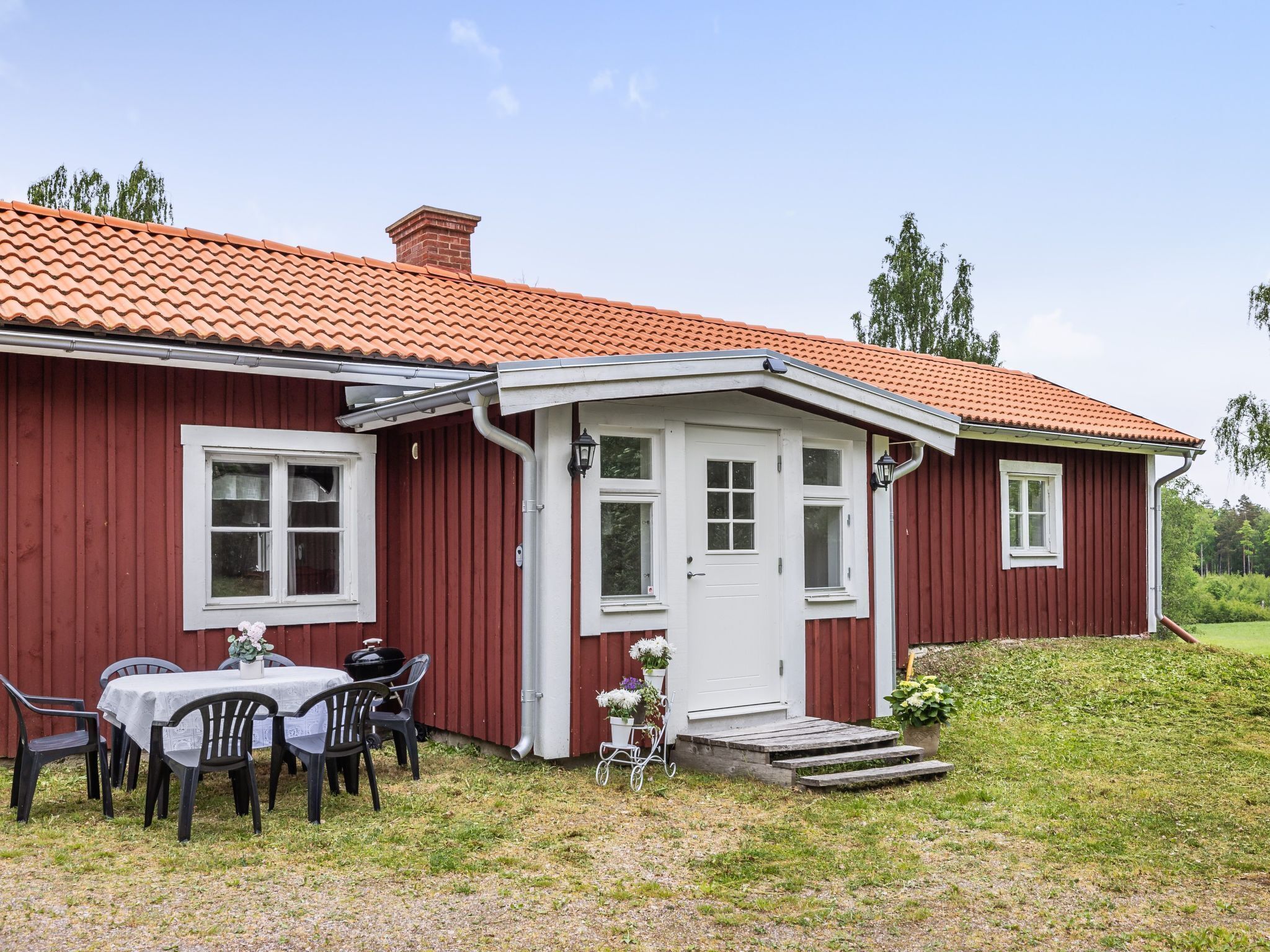 A red wooden house with a white door and windows, a small porch, and a table with chairs outside.