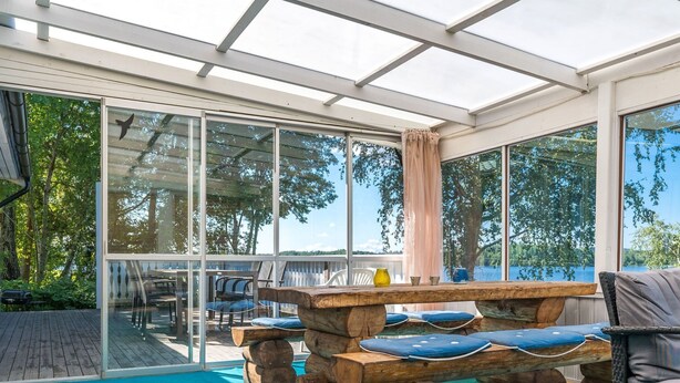 A covered outdoor area with a wooden table and chairs, large windows, and a view of trees and water.