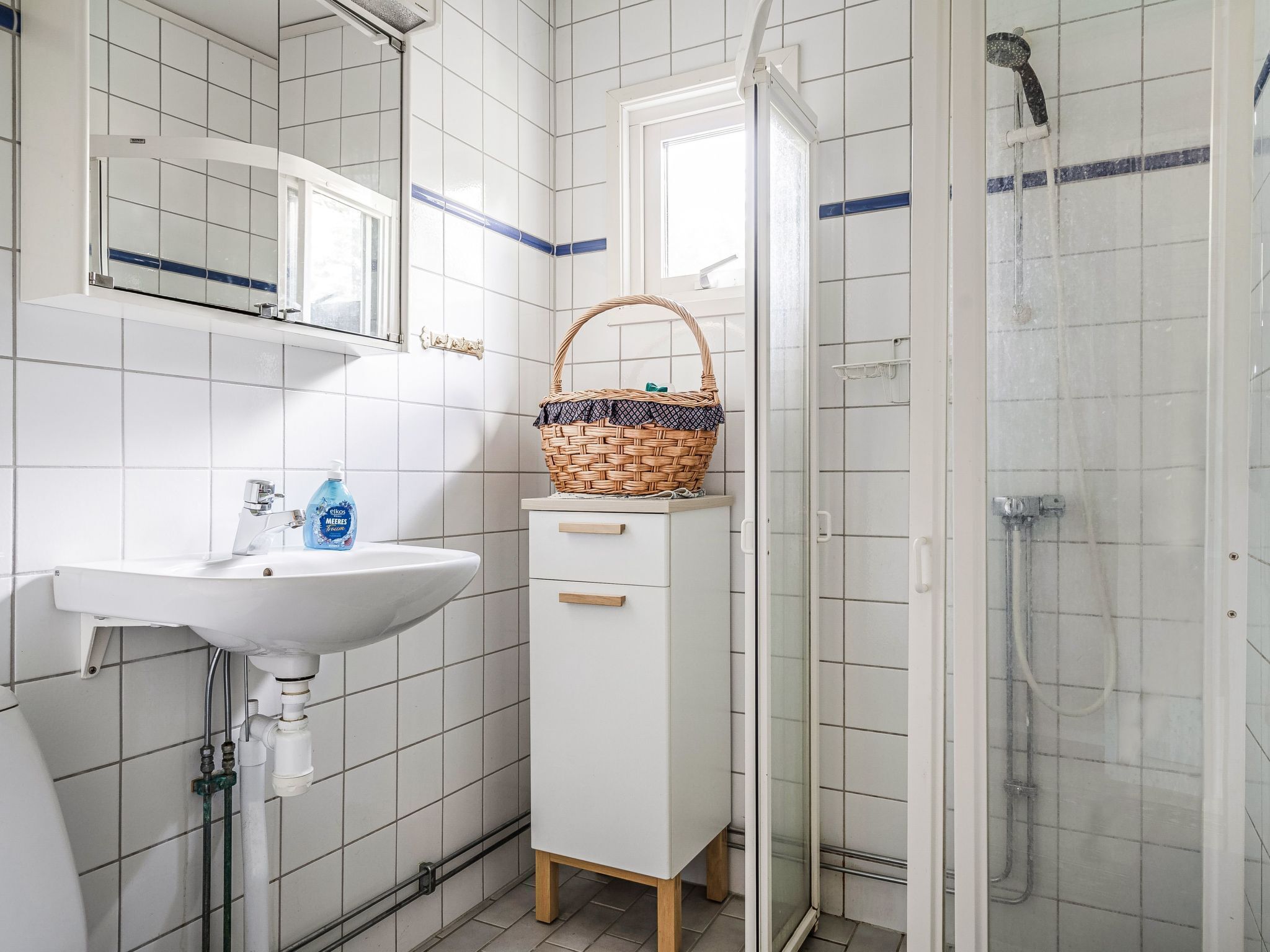 Een badkamer met een witte wastafel, een spiegel, een douche en een mandje op het aanrecht.
