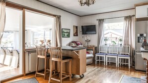 Ein gemütliches Zimmer mit einem Holzesstisch, Stühlen, einer kleinen Küchenzeile und einem großen Fenster mit Vorhängen.