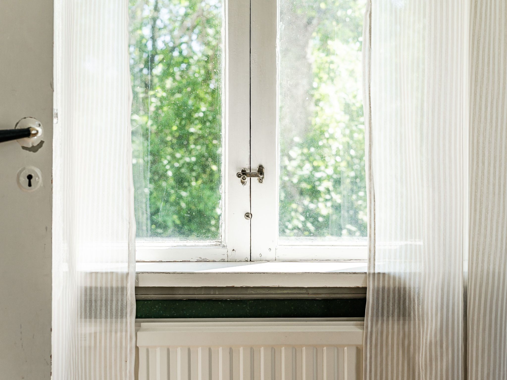 Une porte blanche, dotée d’un trou de serrure et d’un loquet, qui donne sur une fenêtre offrant une vue sur des arbres verdoyants.