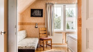 Et soveværelse med en seng, en stol, en radiator, et vindue med gardiner og en lampe monteret på væggen.