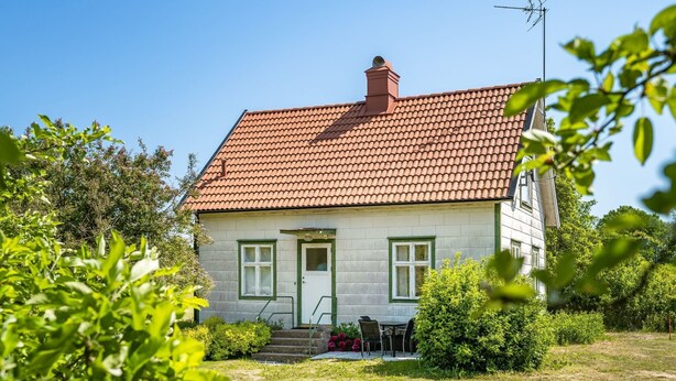 Et hus med røde tegltage, hvide vægge og en grøn dør. En lille have med græs og buske, samt et bord og stole udenfor.