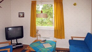 Una stanza con una televisione, due poltrone blu, un tavolo con tovaglia a quadri e una finestra con tende gialle.