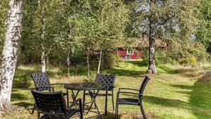 Et udendørs siddeområde med et bord og fire stole, omgivet af træer og en rød bygning i baggrunden.