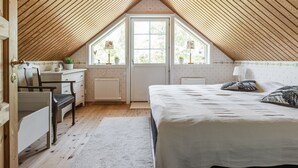Ein gemütliches Schlafzimmer im Dachgeschoss mit schrägen Holzbalken, einem Bett mit weißer Bettdecke, einem Schreibtisch mit Stuhl und einem Fenster mit Blick auf Grünflächen.