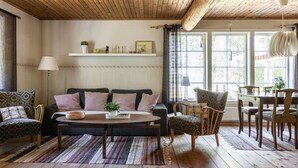 Ein gemütliches Wohnzimmer mit Holzbalkendecke, einer Sofagarnitur, einem Esstisch und einem großen Fenster, das für natürliches Licht sorgt.