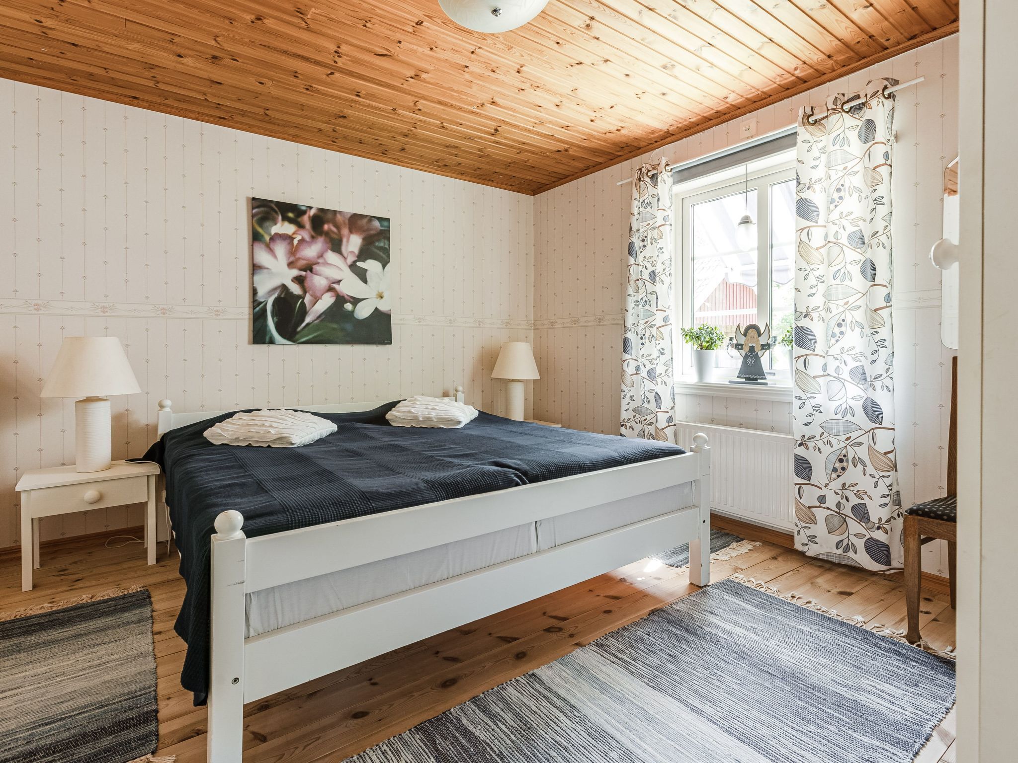 A bedroom with a wooden ceiling, a bed with white headboard, two bedside tables with lamps, a window with floral curtains, and a wall-mounted painting of flowers.