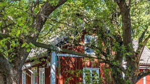 Une maison en bois rouge, dotée d’un porche, entourée d’arbres et d’herbe d’un vert luxuriant.