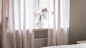 Une pièce avec des rideaux légers, un radiateur blanc et un vase rempli de fleurs posé sur un rebord de fenêtre.