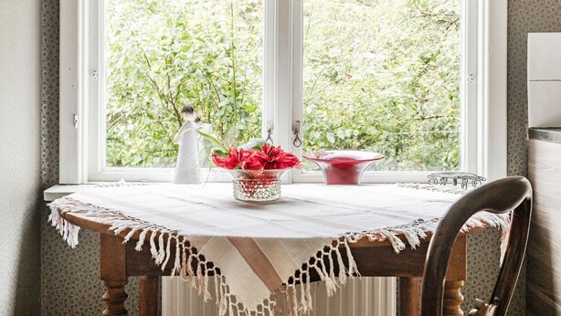 Une table à manger recouverte d’une nappe blanche, un vase avec des fleurs rouges et un bol rouge.