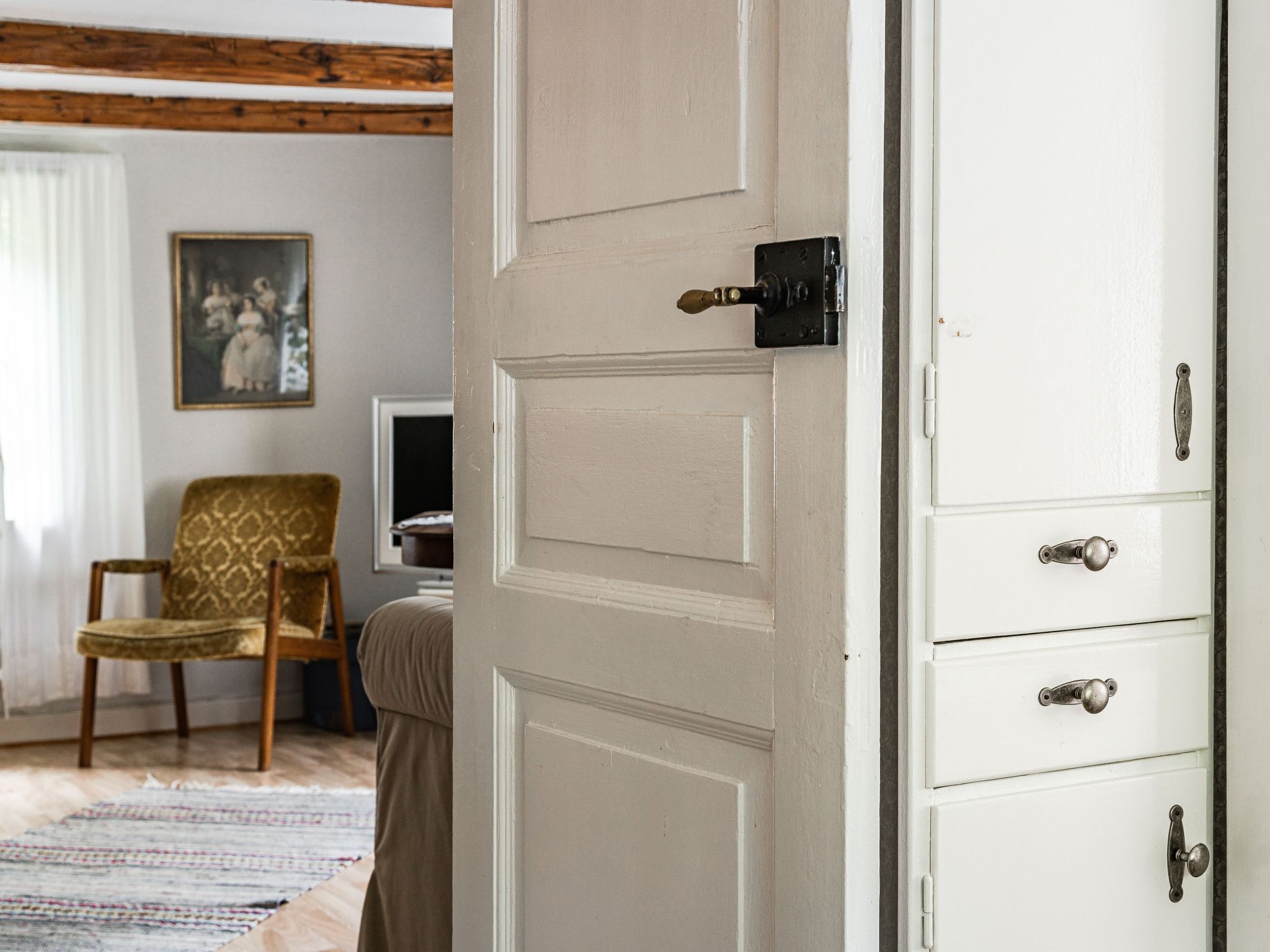 A room with a white door, a wooden chair, a white cabinet with drawers, and a framed picture on the wall.