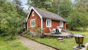 Une maison en bois rouge, avec un toit en pente, un petit porche et un chemin menant à l’entrée.