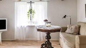 Un salon chaleureux avec un plafond en bois, une table ronde recouverte d’une nappe en dentelle, un canapé beige et un téléviseur posé sur un meuble.