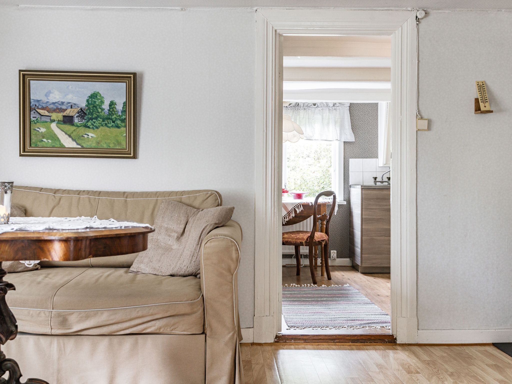 Un salon avec un canapé beige, une table basse en bois et un tableau représentant un paysage. Une porte donne sur une cuisine équipée d’un évier et d’une fenêtre avec un rideau.