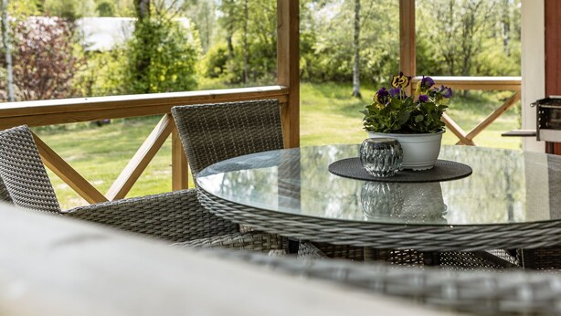 A covered outdoor seating area with a glass table, wicker chairs, and a potted plant with flowers. - Björkebo by Interhome (Ätran)