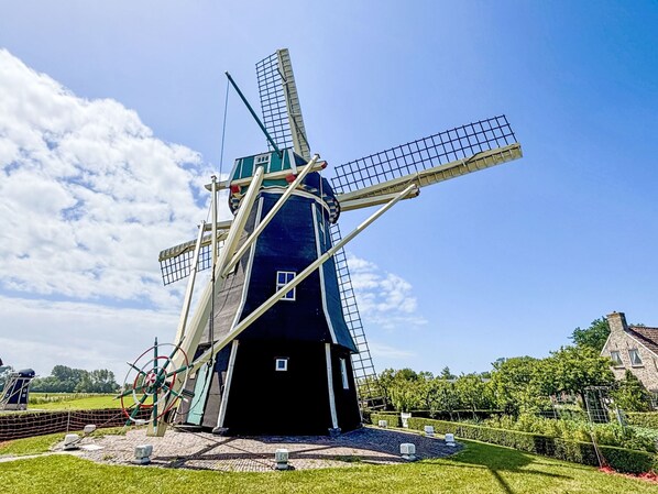 A traditional windmill with a lattice structure, surrounded by a well-maintained garden and a historic building in the background.
