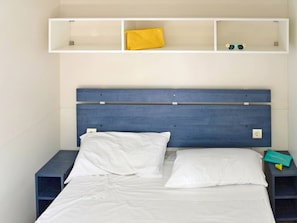 A bed with white linens, a blue headboard, and a shelf above containing a yellow pillow, a pair of sunglasses, and a small item.
