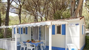 Une petite maison de campagne à un seul étage, à l’extérieur blanc et aux volets bleus, dotée d’un patio couvert avec des chaises blanches et une table dressée pour les repas, le tout entouré d’une végétation luxuriante.