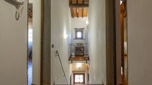 Un couloir étroit, doté de planchers en bois et de murs en pierre, mène à un palier du deuxième étage où se trouve une petite fenêtre et une étagère ornée de fleurs.