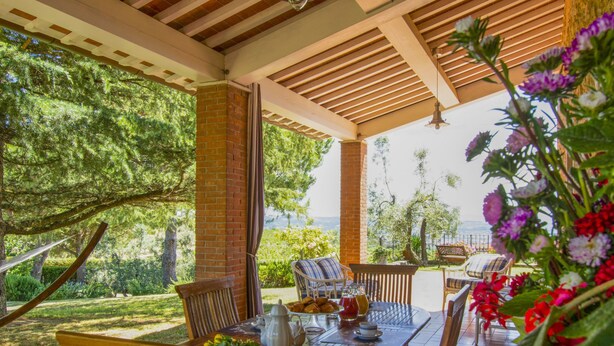 Une terrasse extérieure avec une table en bois dressée pour le petit-déjeuner, entour ée de chaises, et offrant une vue sur un jardin luxuriant et un paysage pittoresque.