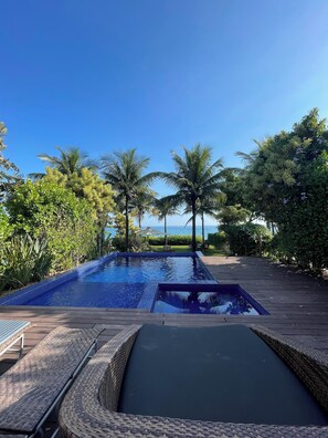 Una piscina rectangular con borde de azulejos, rodeada de exuberante vegetación y palmeras, con una vista despejada del océano al fondo.