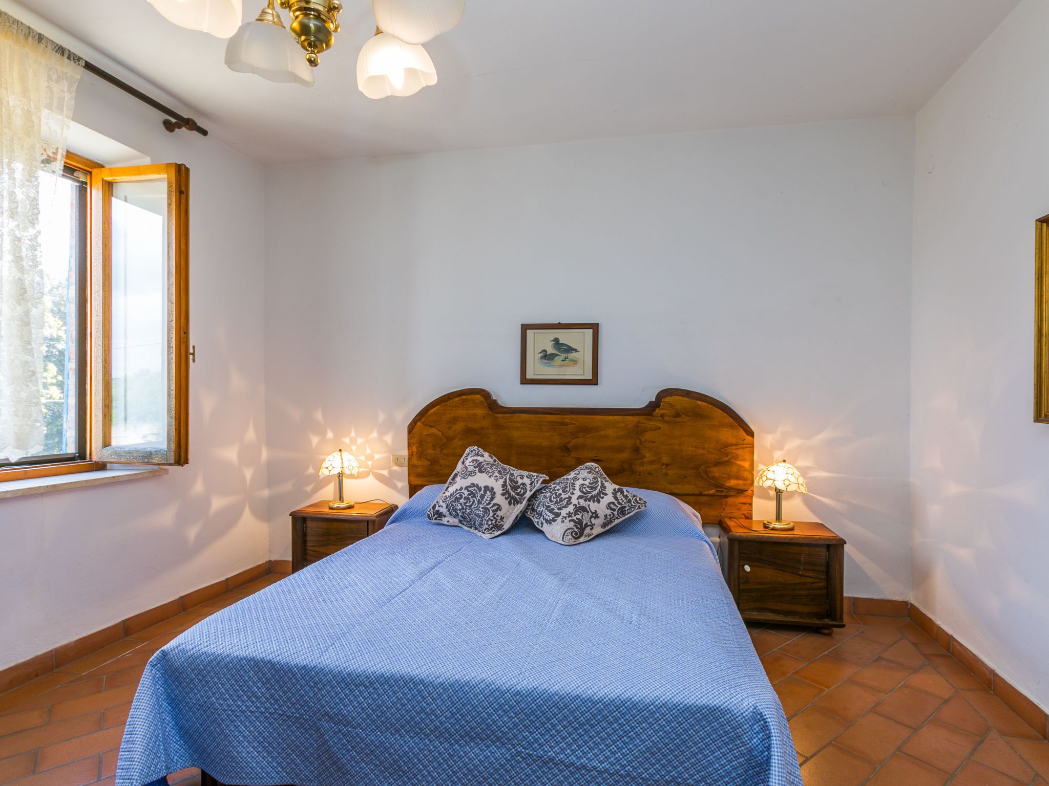 Une chambre à coucher avec une tête de lit en bois, un lit simple recouvert d’une courtepointe bleue, deux lampes de chevet, une fenêtre avec des rideaux et un tableau encadré accroché au mur.