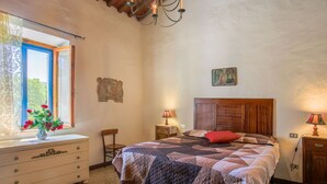 Camera da letto con un letto di legno, un comodino, una cassettiera, una finestra con tende e un lampadario.